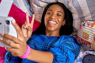 Smiling girl in blue shirt takes a selfie and makes a peace sign while lying on a bed.
