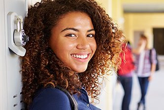 Smiling student with curly hair leans against a locker in a school hallway.