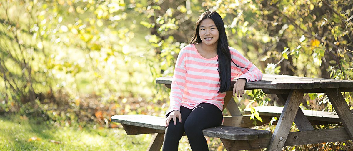 A girl smiles while sitting on a picnic table in a sunlit park with green foliage.