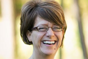 Woman with short brown hair and glasses smiling widely outdoors in natural light.