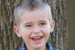 Smiling young boy with short hair stands in front of a tree, wearing a plaid shirt.