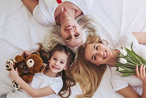 Three generations of smiling women lying on a bed, holding a teddy bear and white tulips.