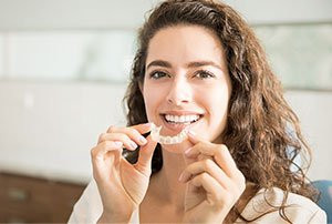 Woman with curly hair smiling and holding a clear dental aligner in her hands.