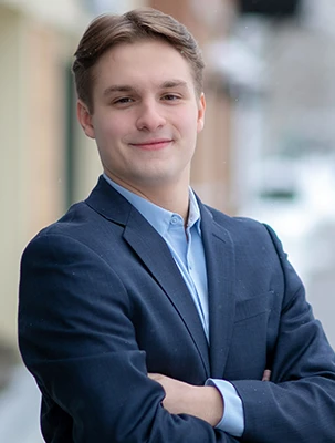 Person in a blue suit smiling with arms crossed, standing on a street with blurred background.